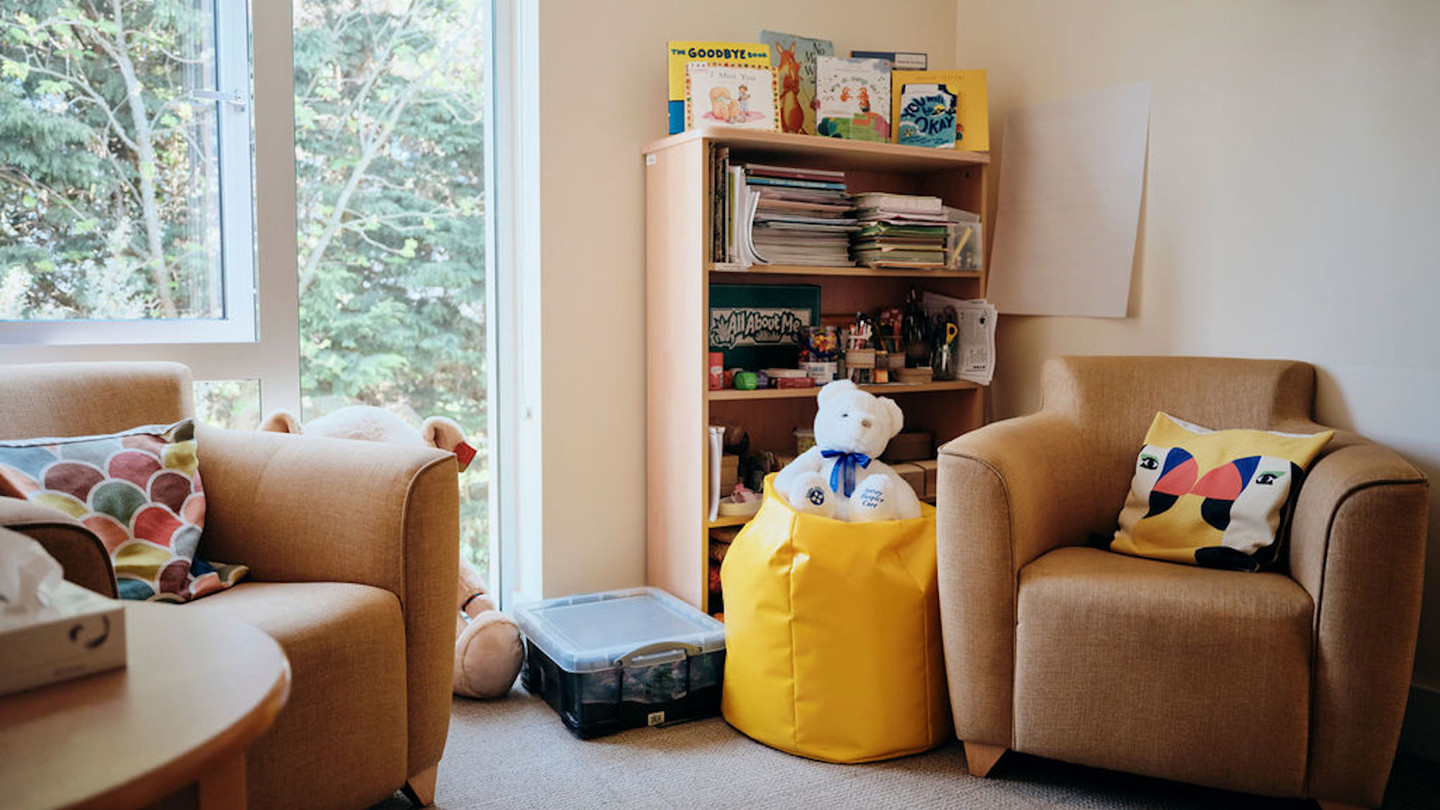 Image of bereavement room, two chairs and a table, with books in the corner and a teddy