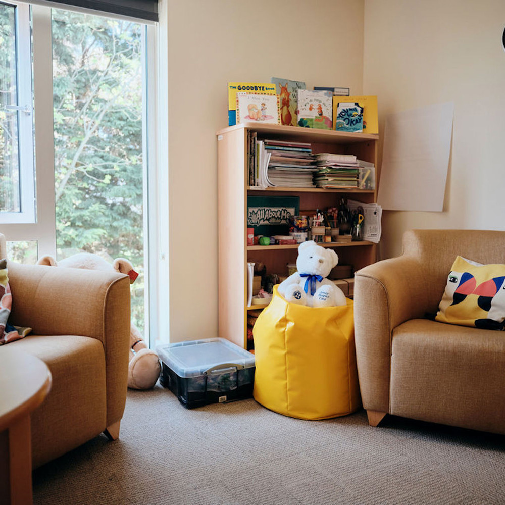Image of bereavement room, two chairs and a table, with books in the corner and a teddy