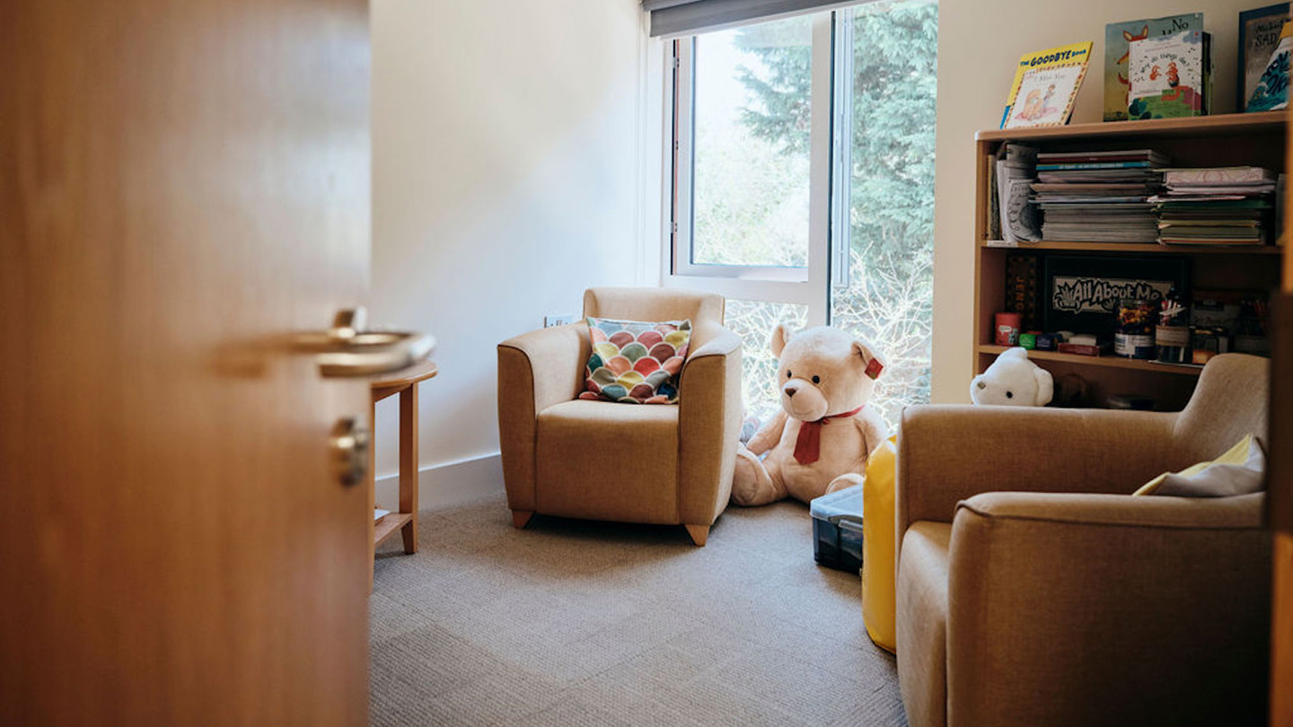 Image of bereavement room with door half open.  Two chairs and a table with large teddy in corner